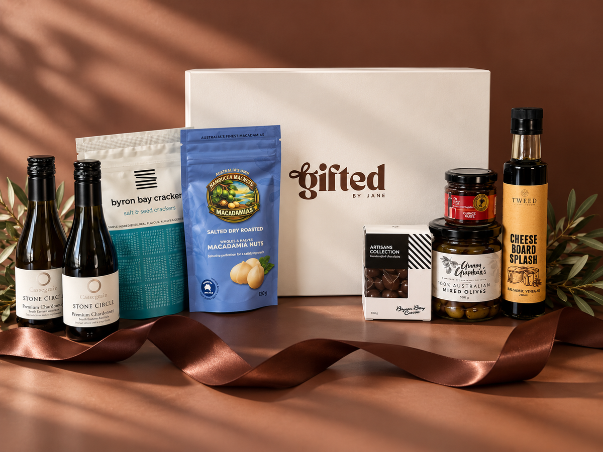 A curated gourmet gift hamper set on a brown surface with a matching brown ribbon. The set includes a large white 'Gifted by Jane' box, a bottle of Cassegrain Wine, a bag of Byron Bay Crackers, a blue bag of Nambucca Macnuts Macadamia nuts, a box of Byron Bay Cacao chocolate, a jar of The Other Chef Quince Paste, a jar of Grumpy Grandma's mixed olives and a bottle of Tweed Real Food Cheeseboard Splash.